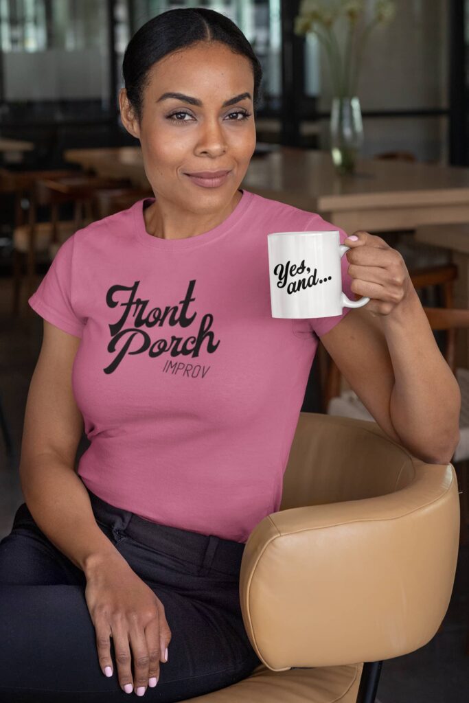 Custom merch for Front Porch Improv by Olive Ridley Studios: A woman wearing a pink t-shirt that features the Front Porch Improv logo printed in black. She is holding a white coffee mug with the phrase "Yes, and..." printed on one side.