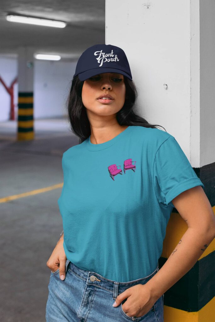 Custom merch for Front Porch Improv by Olive Ridley Studios: A woman wearing a teal "dad tee" featuring pink chairs printed on the left chest. She is also wearing a navy baseball cap with "Front Porch" embroidered in white across the front.