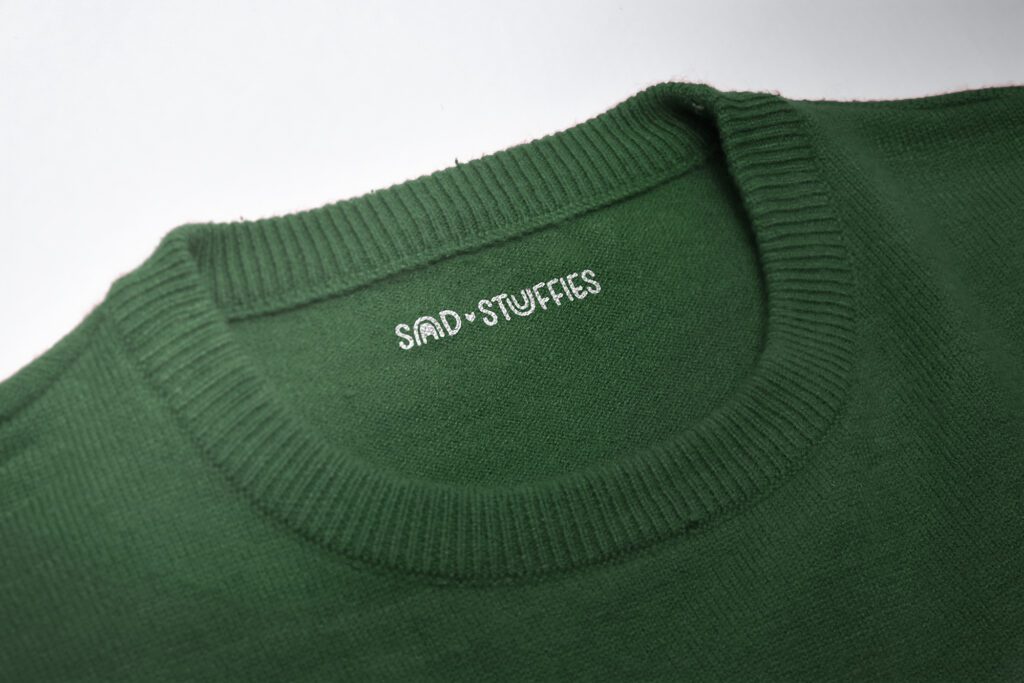 Brand identity design created for Sad Stuffies by Olive Ridley Studios - Secondary logo shown mocked up as a printed tag on the inside of a sweater